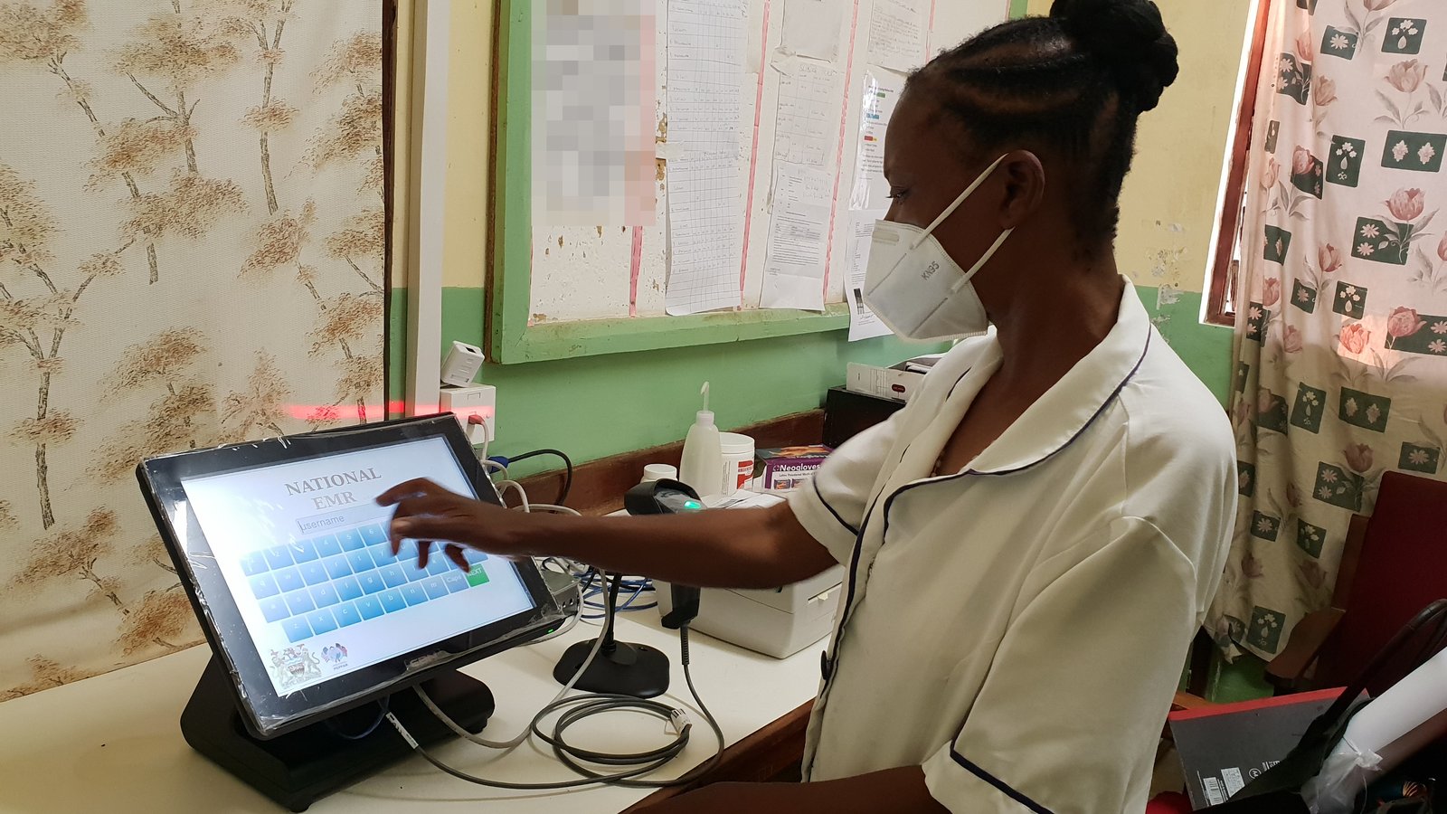 A health worker in a face mask logs in to the National EMR touchscreen at an HIV clinic in Malawi, with a barcode scanner and label printer beside her