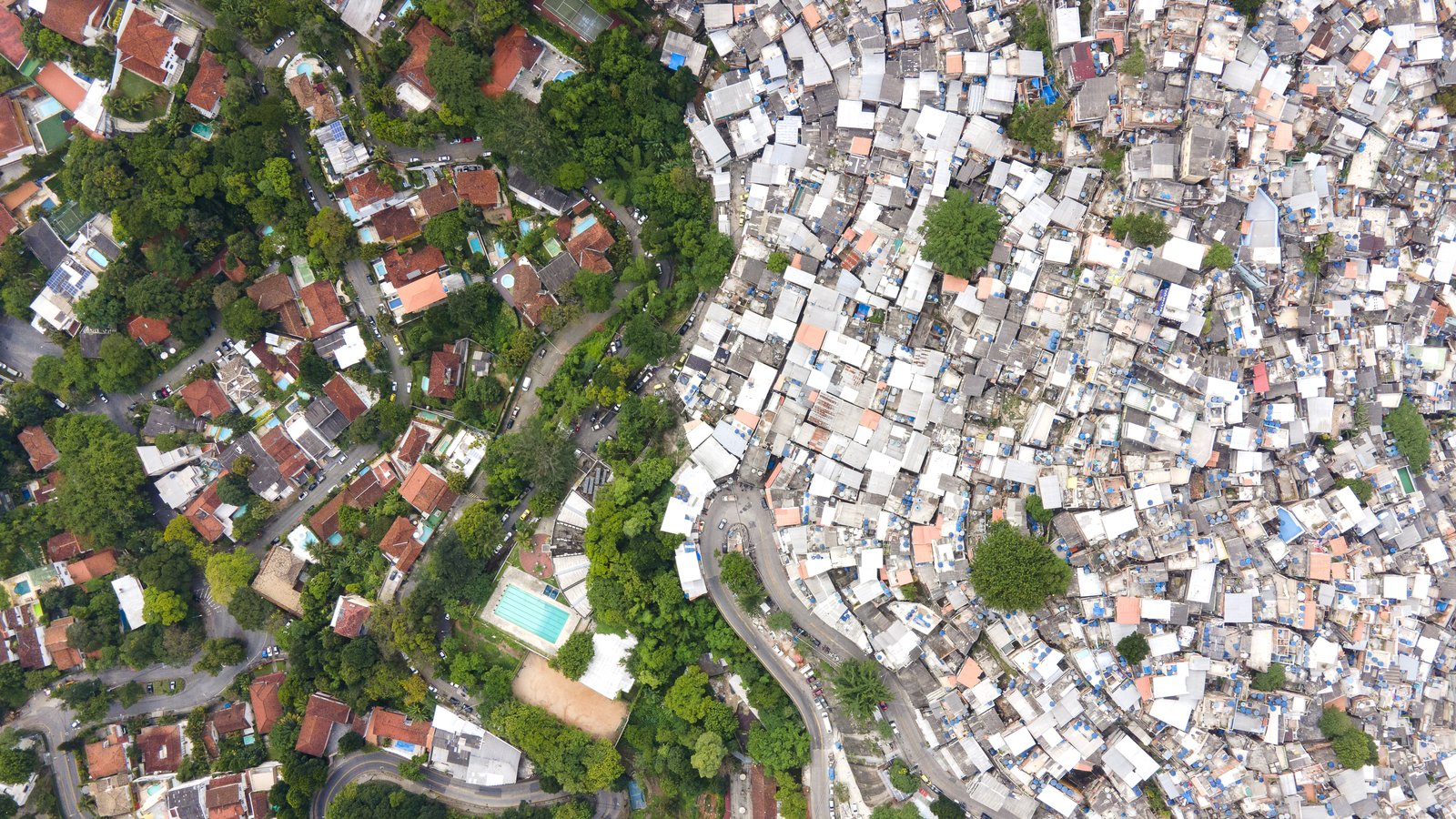Aerial drone view of the border between the Rocinha favela and the Gávea neighborhood in Rio de Janeiro: dense rooftops on one side, houses with pools among trees on the other