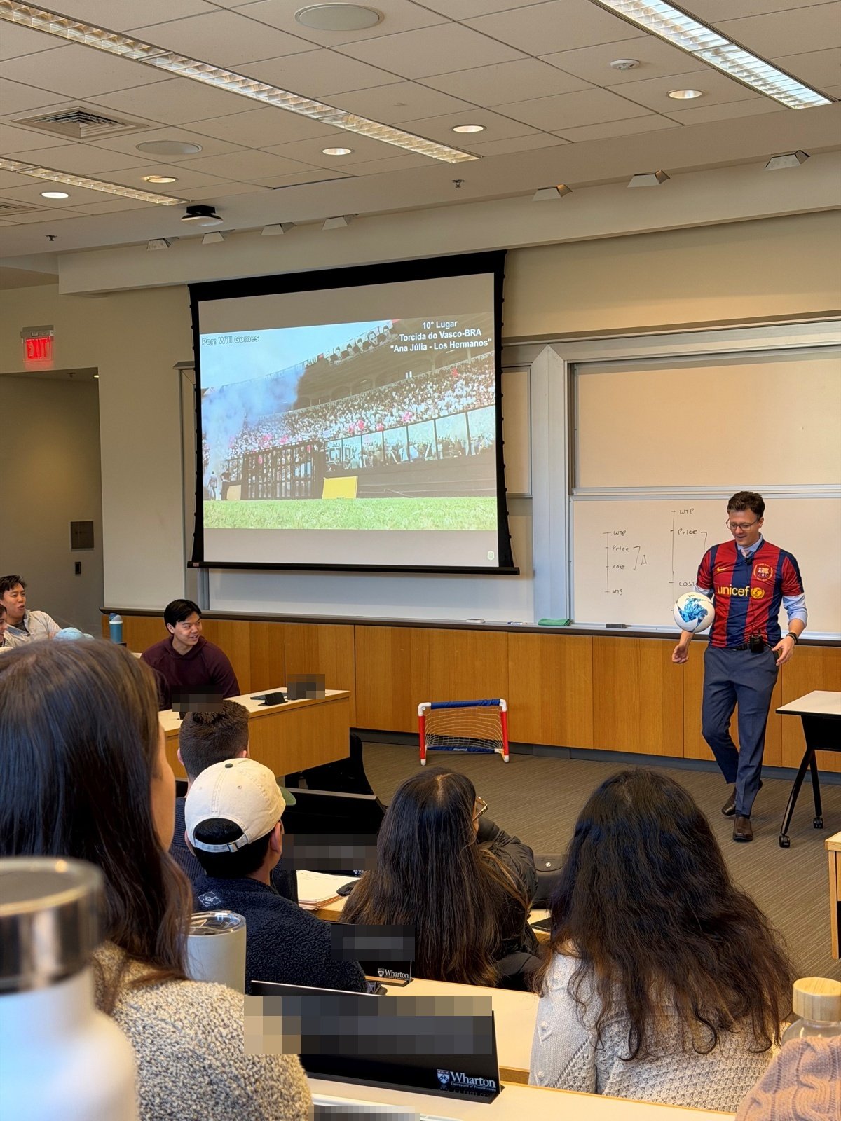 Leandro Pongeluppe at the front of a Wharton classroom wearing a football jersey and juggling a ball, with a slide of a stadium crowd on the screen and students watching from their desks, Fall 2025